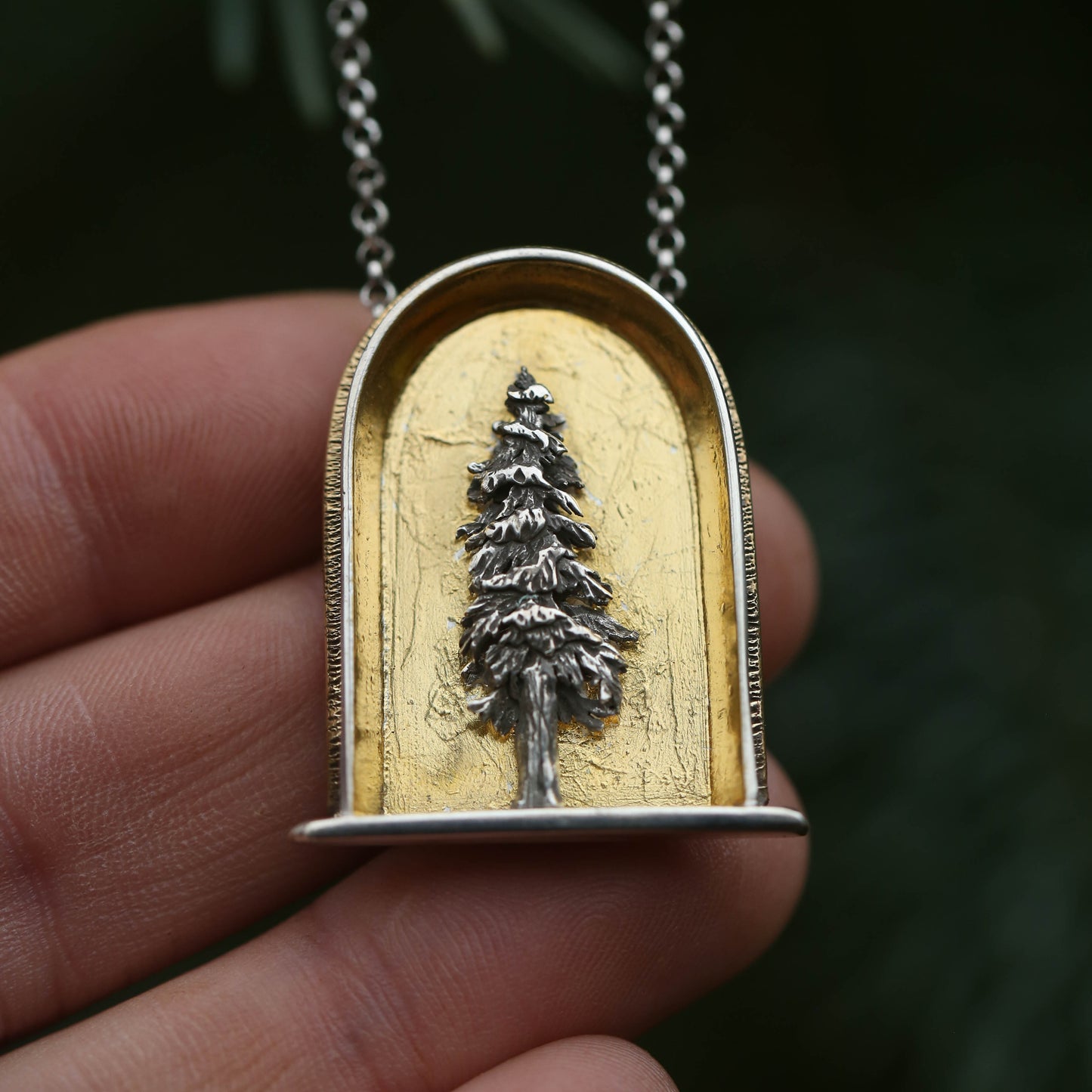 A sterling silver and 24k gold necklace in an arch shape featuring a layered sterlign silver redwood tree with a green background