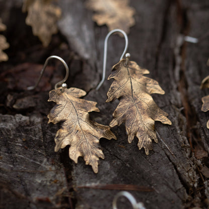 Brass gold colored oak leaf-shaped earrings on a textured wooden surface.