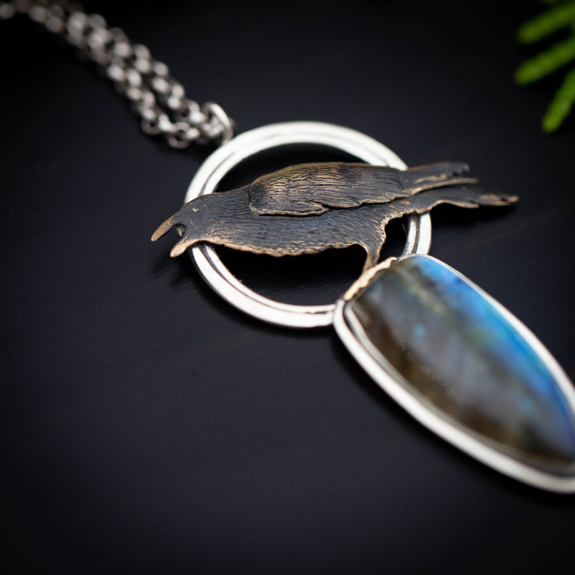 a detail of a necklace featuring a brass raven on an open silver circle utop a tear drop labradorite stone shown on a black background