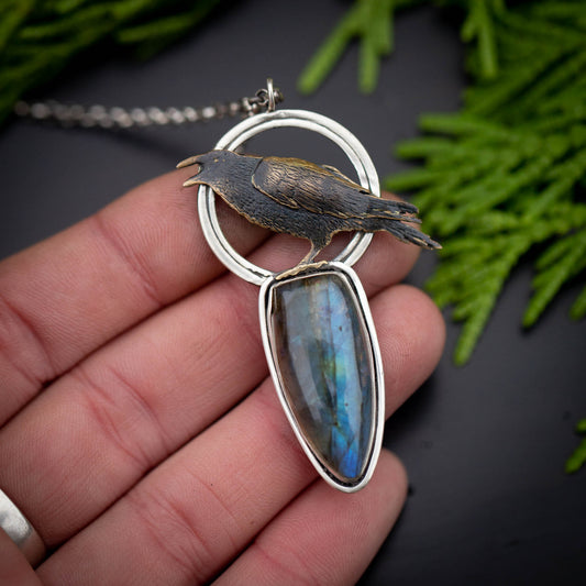 A hand holding a necklace featuring a brass raven on an open silver circle utop a tear drop labradorite stone