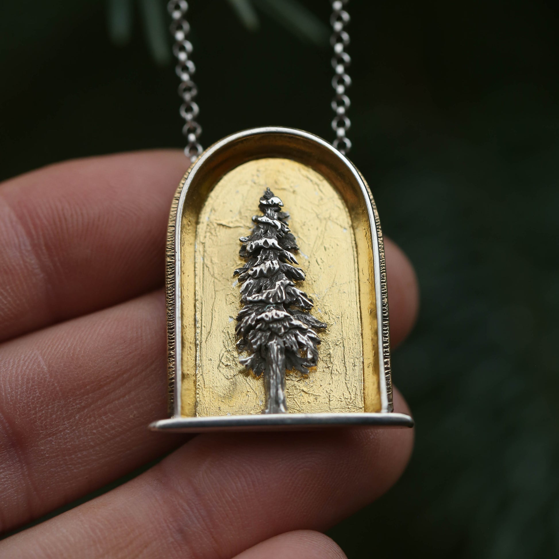 A sterling silver and 24k gold necklace in an arch shape featuring a layered sterlign silver redwood tree with a green background